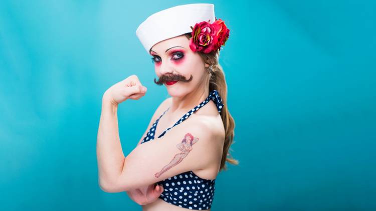 A woman with a moustache in a polka dot bikini top and sailors hat flexes her guns