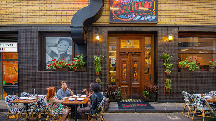 People dining outside on the street at a table sitting on chairs.