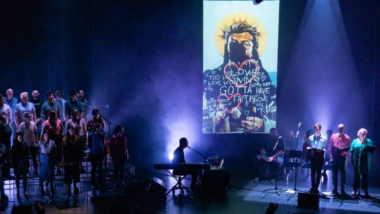 A performance of The Rise and Fall of Saint George, with a choir on stage, Paul Mac playing keyboard, and a projected mural of George Michael 