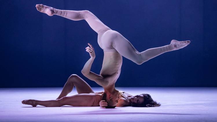 A topless dancer on his head, legs akimbo, with a female dancer lying on the floor behind him 