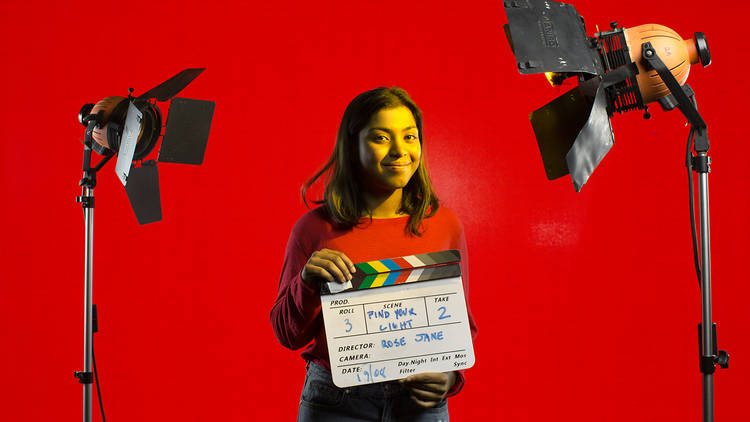 A young person smiles holding a clapperboard with a red background and professional lights