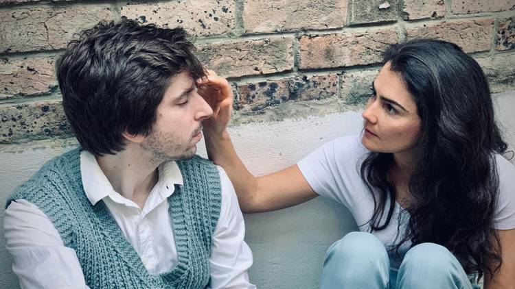 A woman with long dark hair touches the forehead of a man in a pale blu cardigan