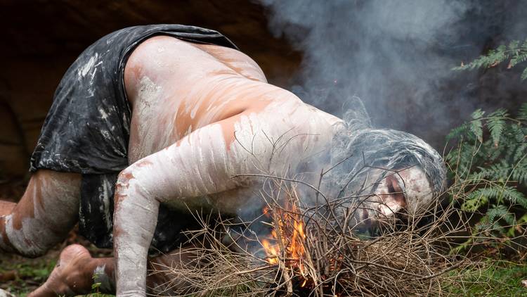 An Indigenous man blows on a fire in ferny bush