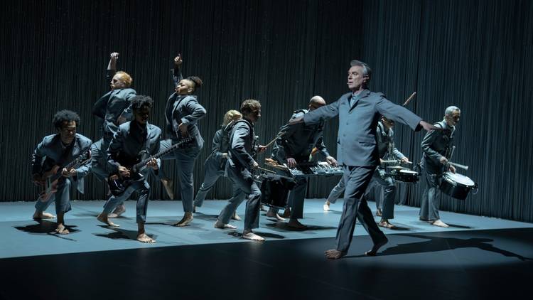 David Byrne in grey suit dances barefoot with his diverse American Utopia bnad 