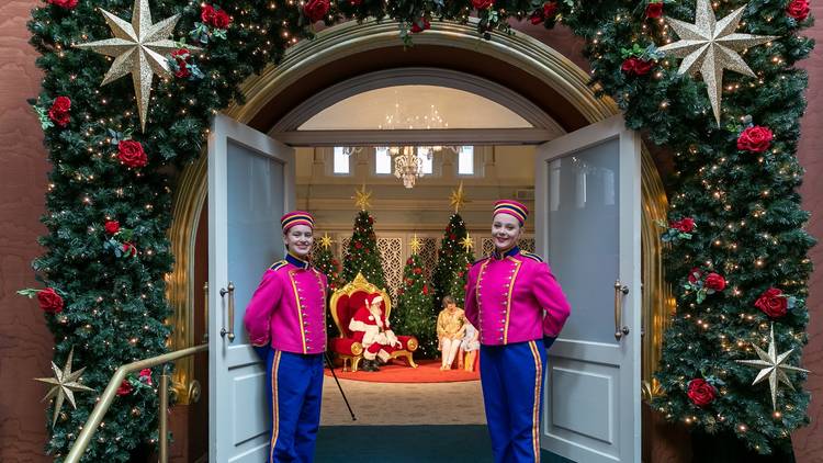 Two people in pink uniforms stand at door to room where Santa sits on throne