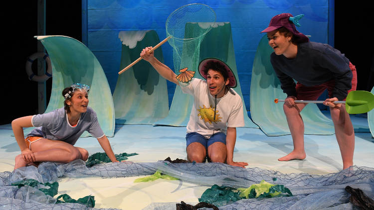 Three performers on stage in front of big curved wave props and surrounded by seaside ephemera