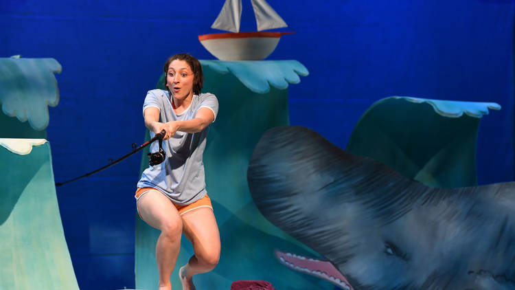 A performer pretends to fish from a deck while two-dimensional whales and ships pass by in the background set