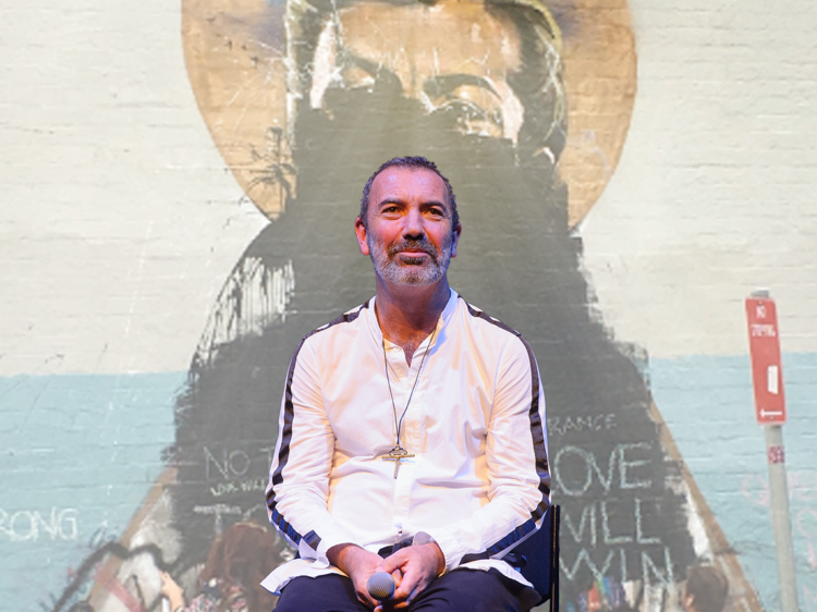 Paul Mac sits on stage, he is superimposed onto an image of a defaced mural.