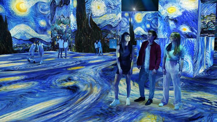 Three people standing and looking around in awe at 'The Starry Night' projected on the walls