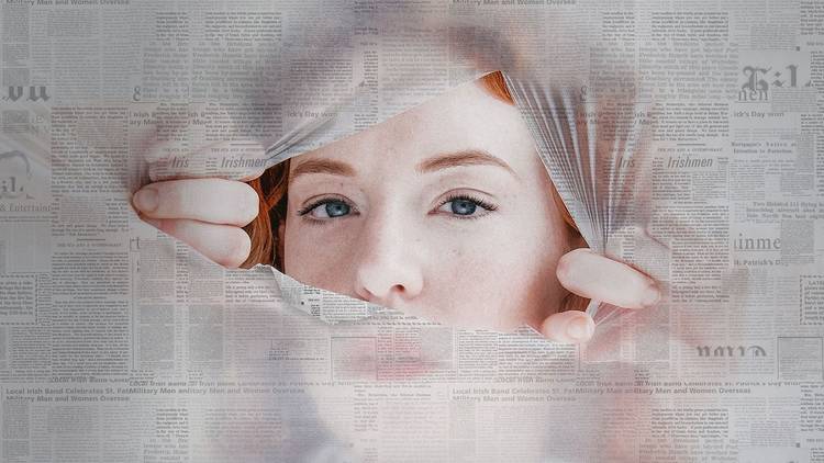 A red-headed woman peering through a torn hole in a newspaper