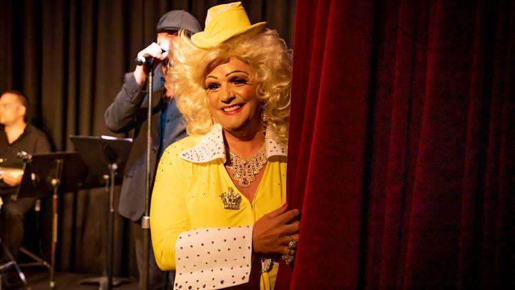 A performer dressed as Dolly Parton smiles from half behind a red velvet curtain