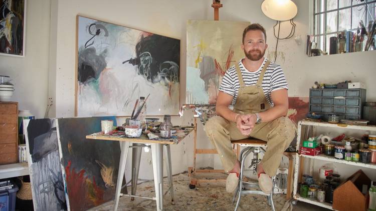 Tim Draxl in a stripey T and painters overalls in his studios surrounded by oil paintings
