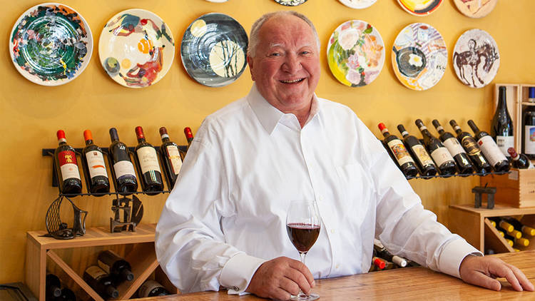 Lucio's owner in a white shirt with a glass of red wine in front of a sunflower yellow wall with painted plate art
