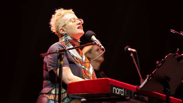 Katie Noonan with short bleached blonde hair in a grey shirt playing a red Korg keyboard