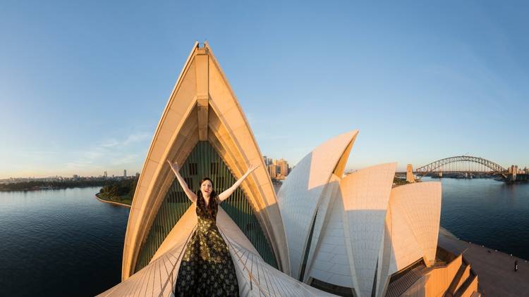 A woman in a fancy dress,  arms outsretched in front of the Opera House 