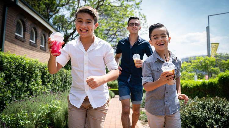 Three young people run through a garden holding colourful drinks and laughing