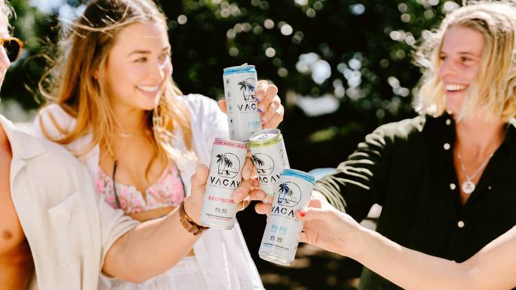 A group of people cheers together cans of hard seltzer in the sunshine.