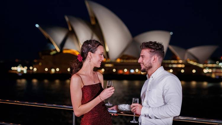 Man and woman drink Champagne on deck of boat, Sydney Opera House is behind them