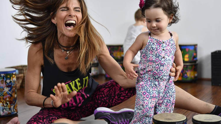 A person laughs with their hair swinging as they play a small set of bongoes. There is also a smiling infant in colourful overalls.