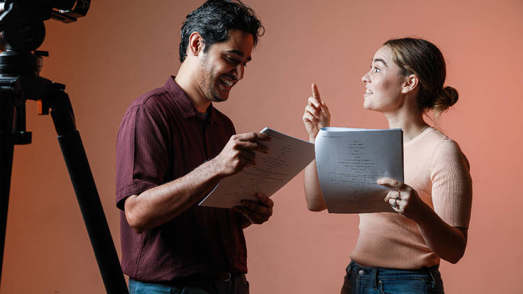 Two people act out a scene, scripts in hand, in front of a pink background. A camera is pointed towards them in the foreground.