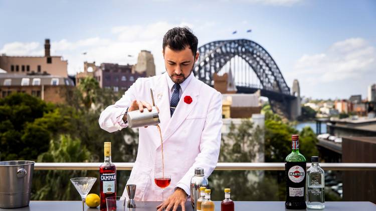 A Mayeb Sammy bartender in s awhite jackets, tie and red carnation mixes a cocktail on the MCA Sculpture terrace with the Harbour Bridge in the background