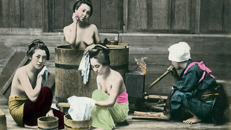 Four people in different stages of undress wash and bath themselves around or in a large wooden barrel bath.