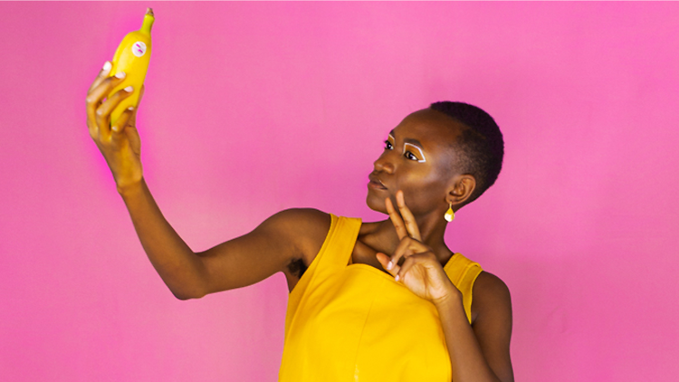 Moreblessing Maturure in a yellow dress holding a banana against a pink backdrop 