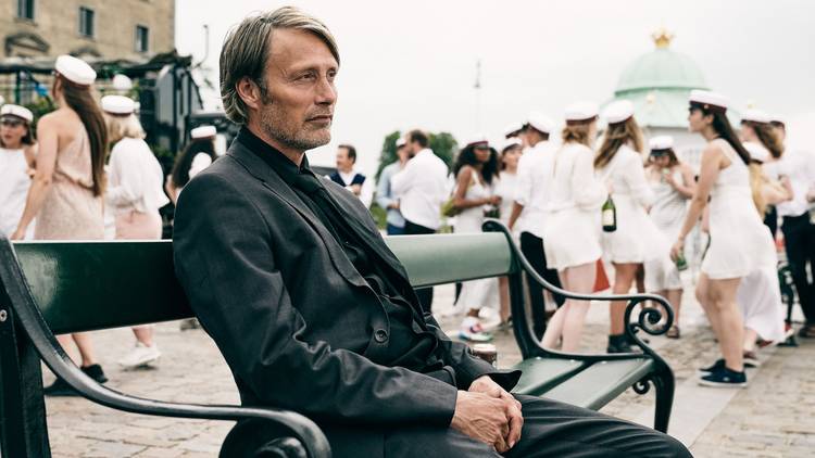 Mads MIkkeslen in a dark suit sits on a bench with students in sailor outfits in the background