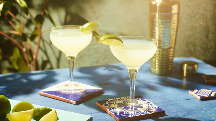 Two Margaritas sit on tile coasters in the sunshine. Both have wedges of lime and are a sunny yellow colour. 