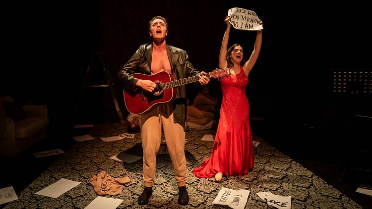 A man wearing a leather jacket plays guitar while a woman in a long red dress holds up a sign featuring lyrics from The Goo Goo Dolls 'Iris'