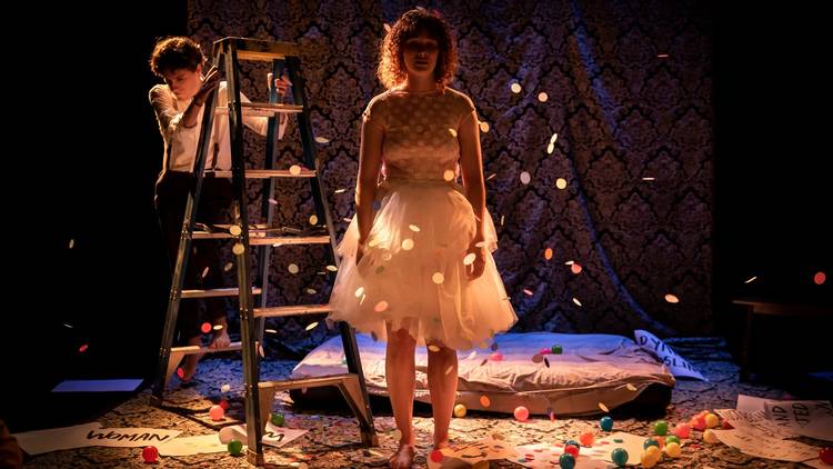 A woman wearing a white tulle dress is heavily backlit so that you can't see her face. A person climbs down a ladder behind her and confetti rains down on them both