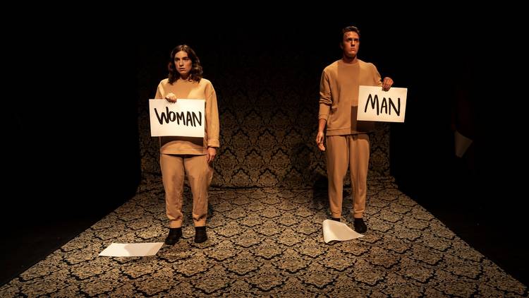 A man and a woman stand expressionless. Both are wearing beige tracksuits and holding up signs that say 'man' and 'woman'
