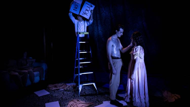 A shirtless man touches the cheek of a woman wearing a long dress. The whole stage is blue-lit and a person stands on a ladder behind the couple, holding signs that say 'not' and 'you'