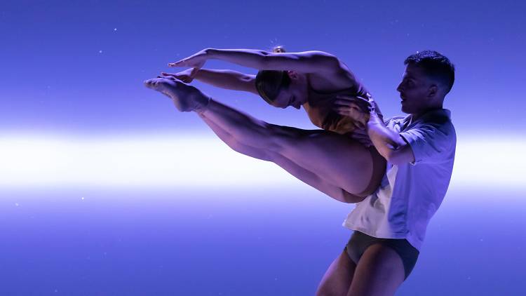 A male dancer holds aloft a woman in a C-formation against a purple backdrop with a ray of white light 
