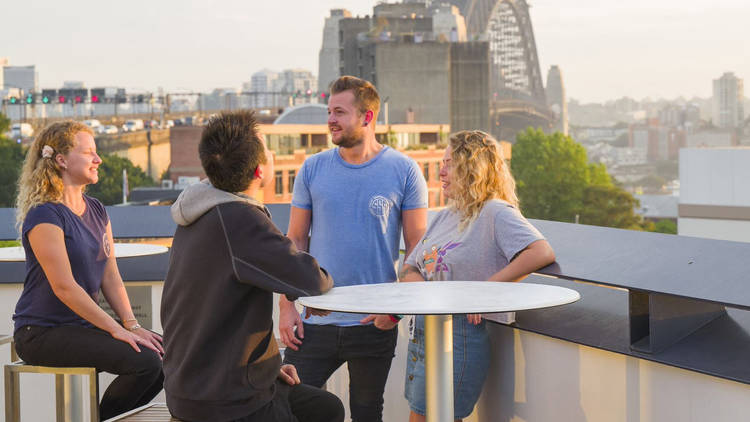 Four people sitting on a balcony as the sun sets. The harbour bridge is in the background. They are on the roof of the YHA Sydney Harbour.