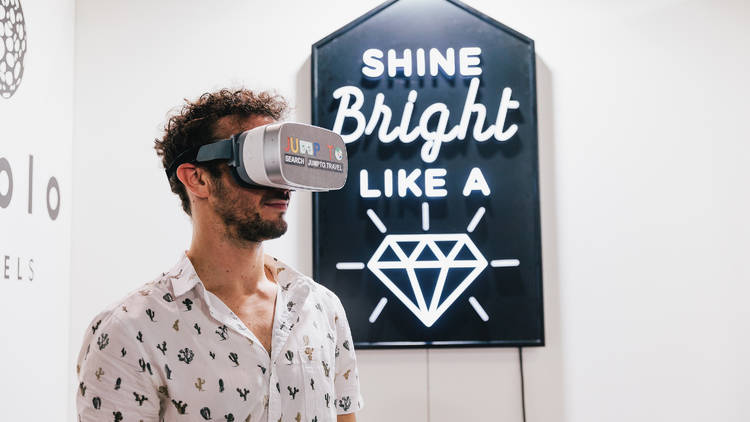 A person wearing a VR headset stands in front of a neon artwork that says "Shine bright like a diamond"