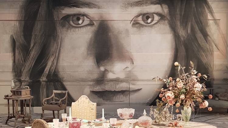 An installation featuring an old fashion table setting; in the background is a realistic mural of a woman's face