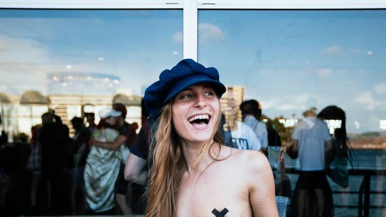 Tilly Lawless smiles at an event and looks away from the camera. She wears a hat and black tape crosses over her bare chest.