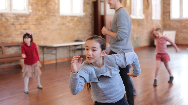 Three kids moving in different postures with a central adult tutor in a wood floor studio space.