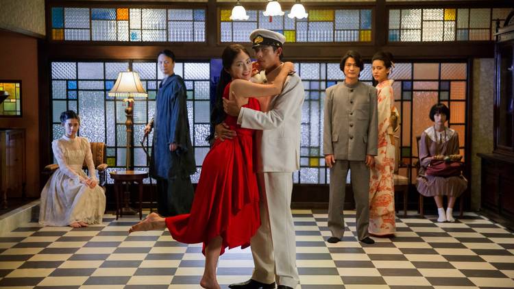 A couple dance on a black and white tiled floor, him in navy uniform, her in a red dress, onlookers witness