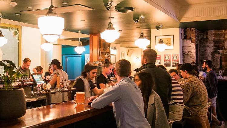 A group of people mill at golden-lit bar. Three bartenders serve the customers. The Unicorn Hotel in Paddington is the location.