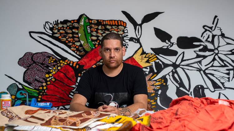 Tony Albert pictured in his studio in front of fabrics and his art on the wall behind him 