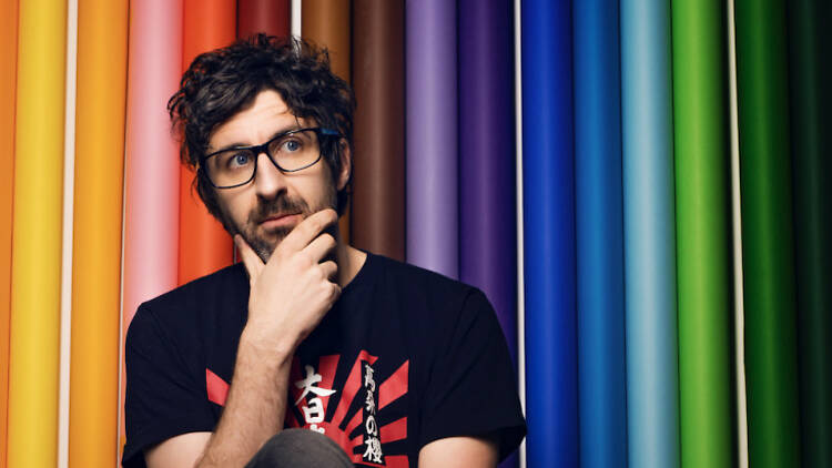 Comedian Mark Watson strokes his bearded chin pensively in front of a rainbow background.