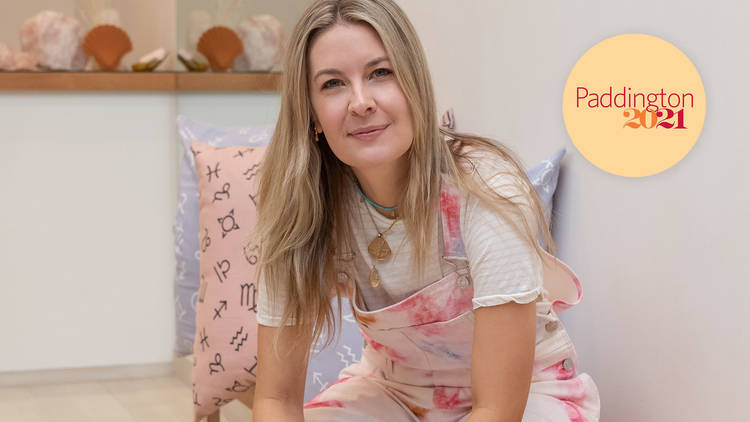 A person wears pastel, tie-dyed overalls on a wood bench with a pink cushion and terracotta and pastel shell sculptures in the background