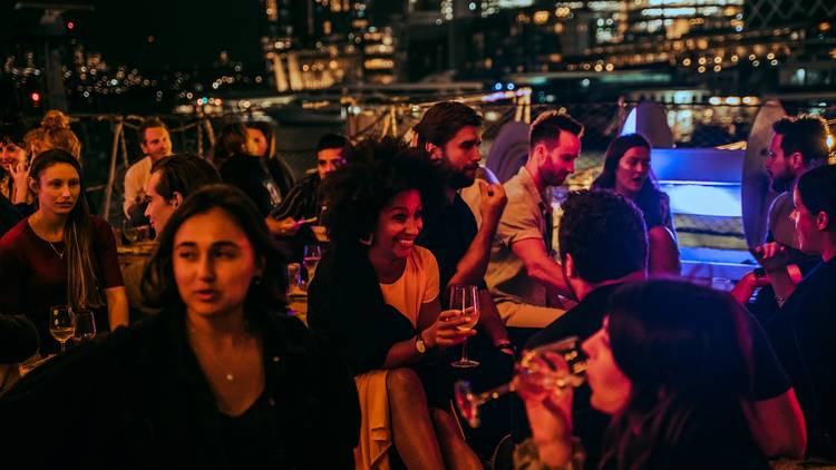 People are seated and drinking on deck of ship with city in background at night