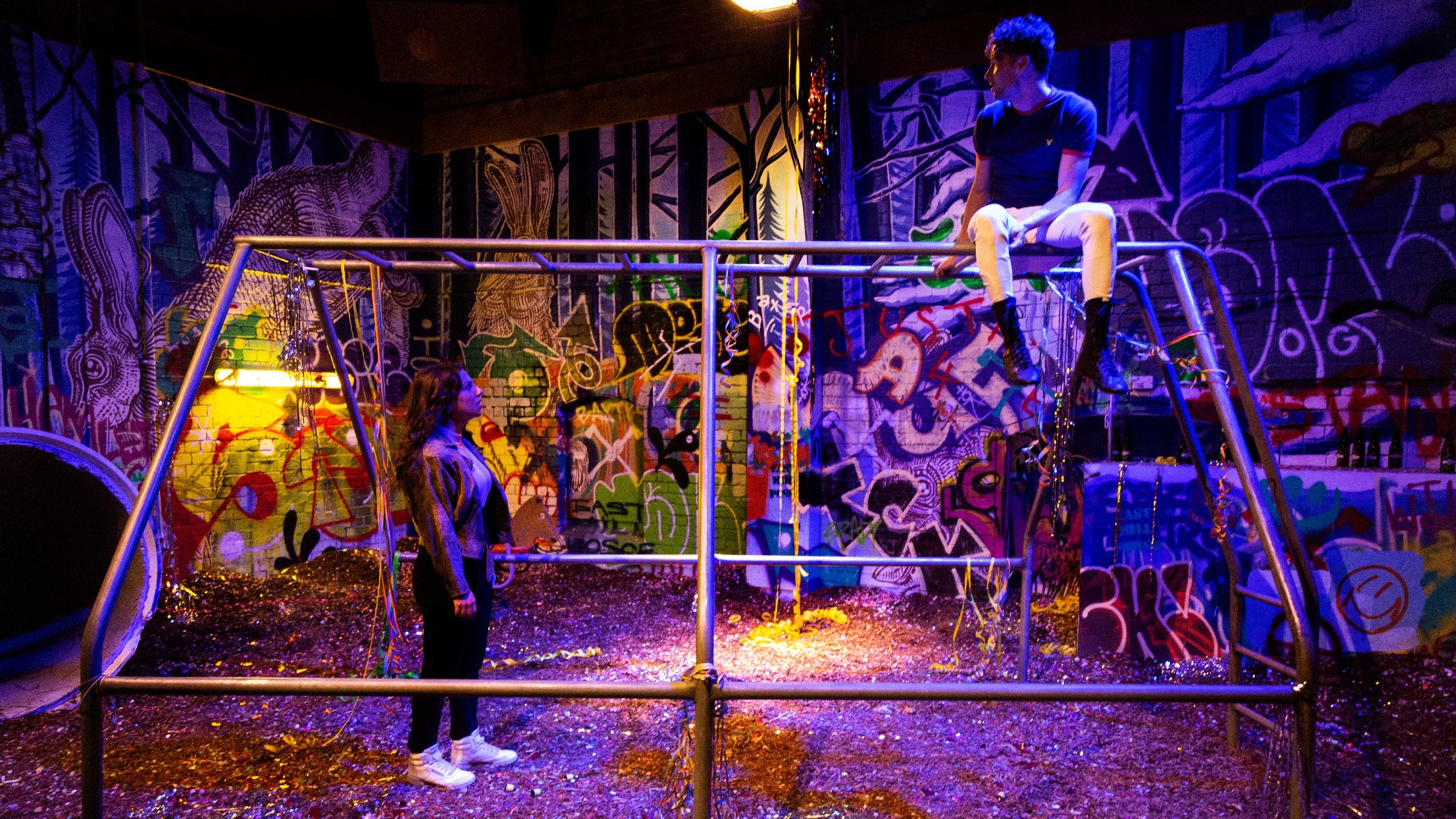 A man sits on a jungle gym in an industrial, heavily grafittied space. He looks down at a woman who is peering up at him.