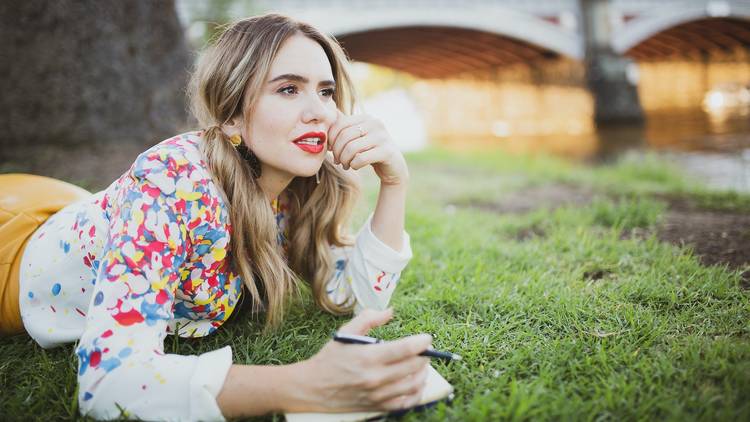 A woman with her hair in pigtails lies on the grass writing notes and looking into the distance