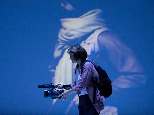 Stop Girl, Belvoir 2021 Sheridan Harbridge, as war reporter suzie, carries a hand held camera, with a projection of a scarf-wearing woman behind her