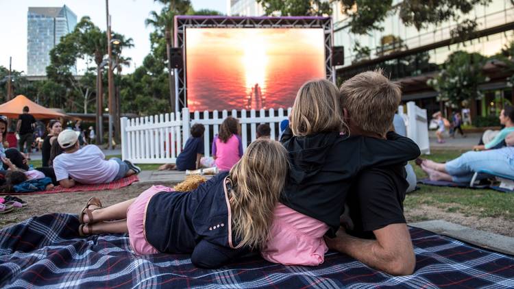 Family cuddled on picnic blanket watching outdoor movie.