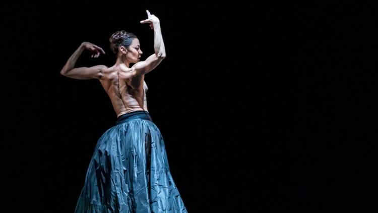 A woman in a ruffled silk blue skirt, bare back, holds her arms aloft dramatically